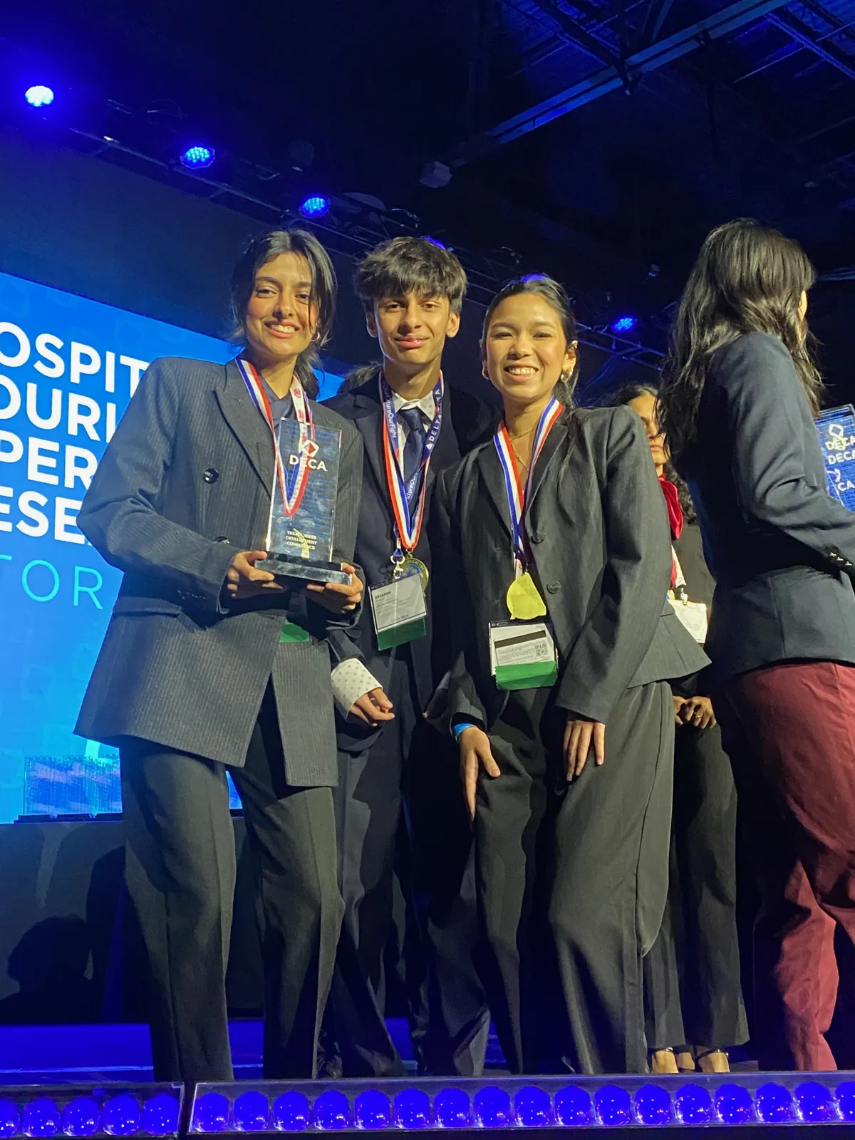 Devansh and two teammates on the Texas DECA stage with medals and a trophy, Hospitality and Tourism Operations Research on the screen behind them