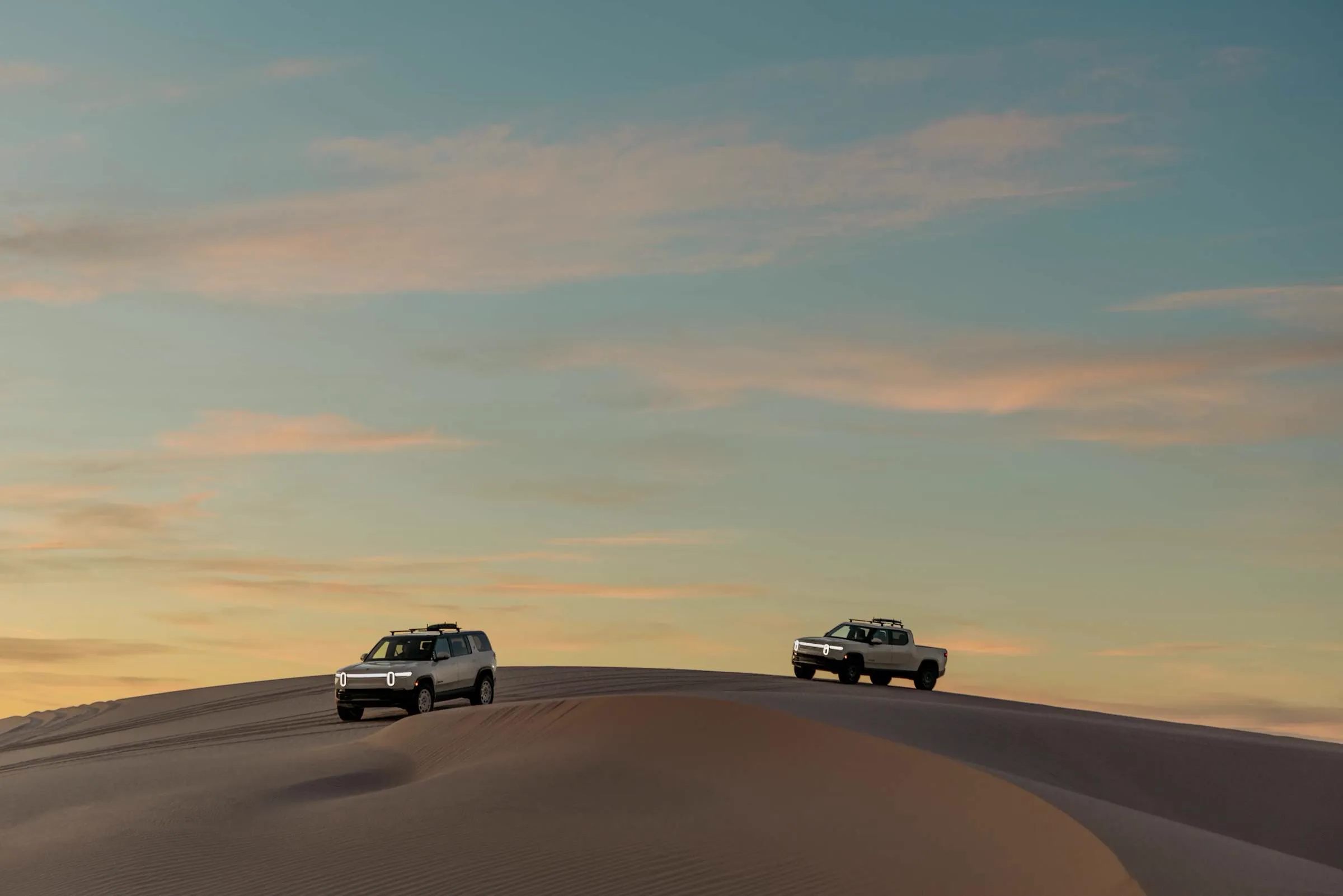 Two Rivian vehicles crossing desert dunes at dusk